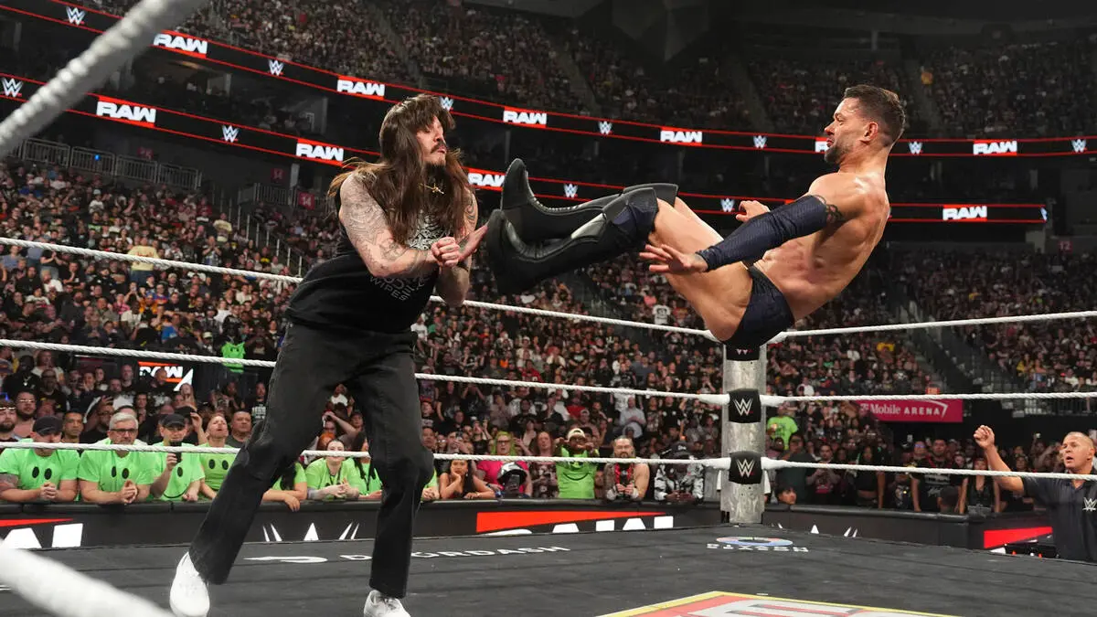 Two WWE wrestlers clash in the ring: one with long hair and a beard braces while the other delivers a high kick from the ropes toward his opponent.