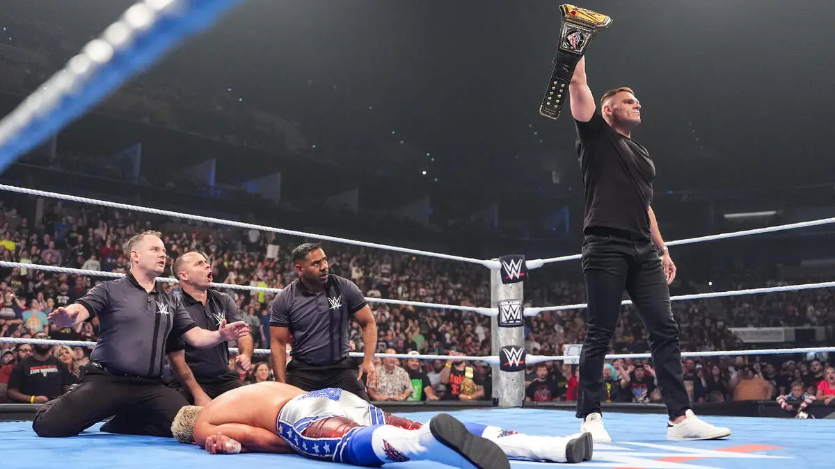 Wrestler in black raises a championship belt above the ring as three teammates kneel beside a fallen opponent on the mat.
