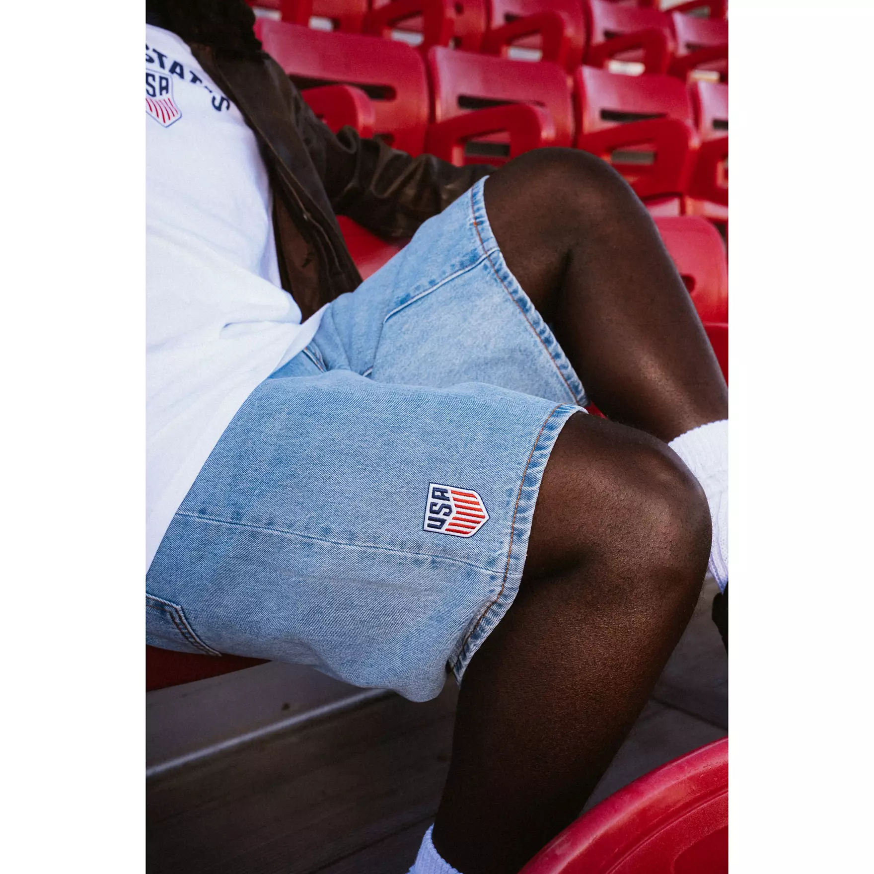 Person seated in red stadium seats wearing light denim shorts with a USA patch visible on the sleeve of a white shirt.