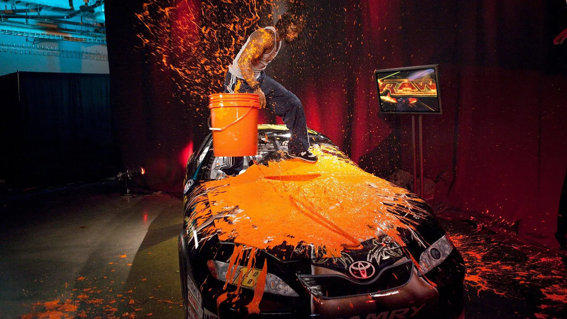 Man stands on a car coated in bright orange paint, bucket in hand, performing a paint-splash stunt indoors.