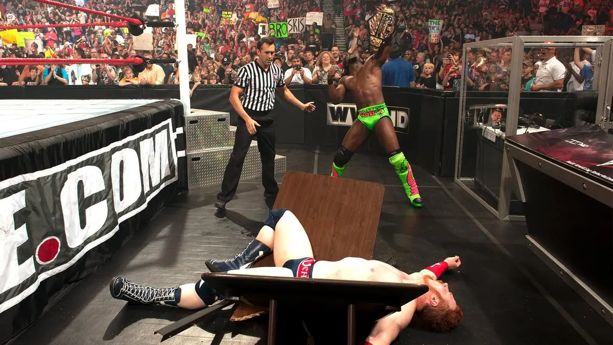 WWE-style wrestling ring scene: a wrestler holds a title belt while a referee looks on; another wrestler lies on the floor under a toppled table as the crowd cheers in the background.
