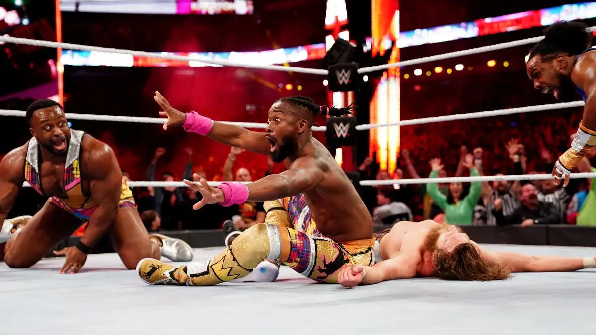 WWE ring moment: a muscular wrestler in colorful tights slides on the mat with arms outstretched as teammates react in the background and a fallen opponent lies nearby, crowd cheering in the arena.