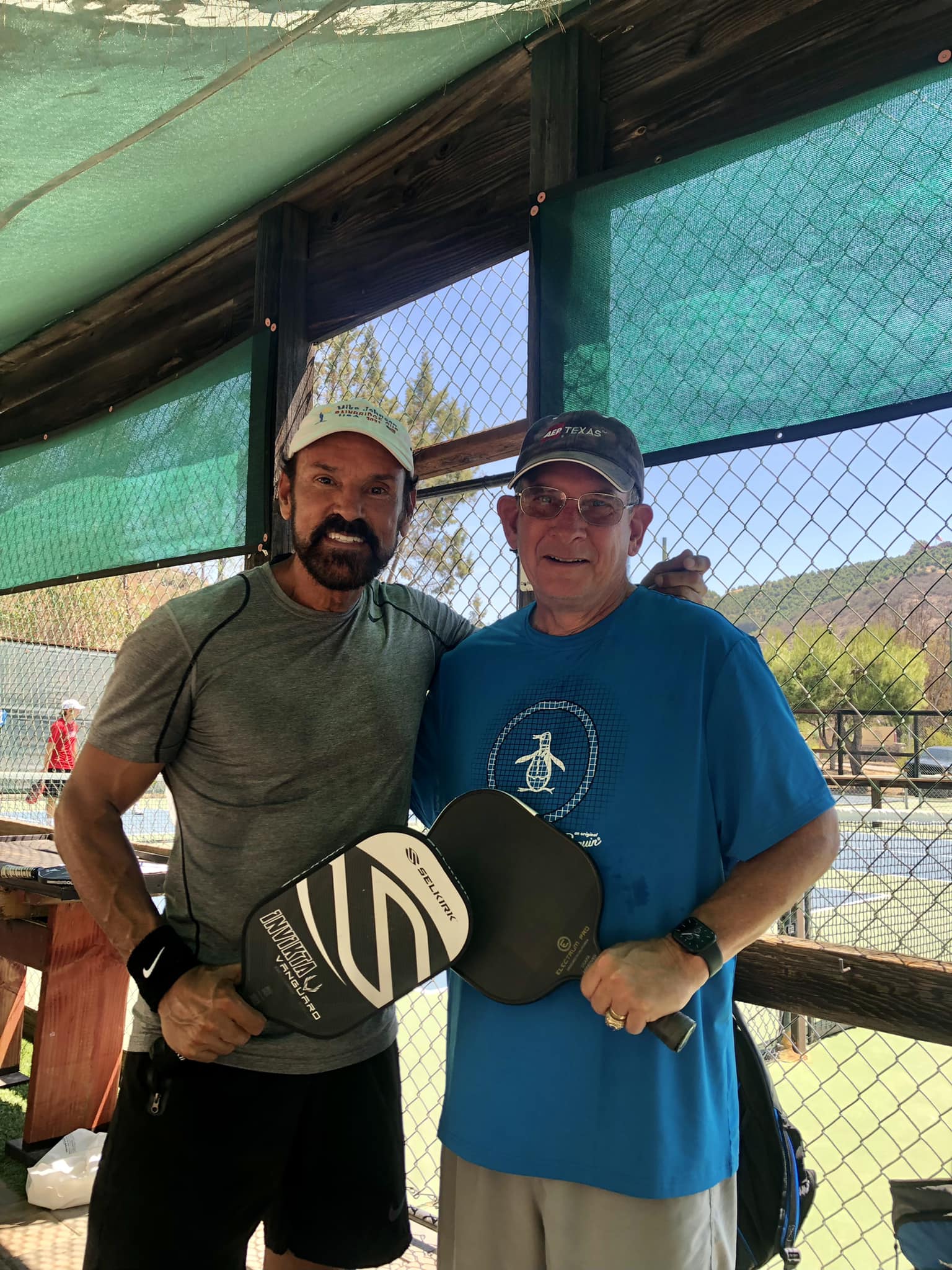 Mike-johnson - the game Two men pose together at a pickleball court, each holding a paddle and smiling at the camera outside under a green canopy.