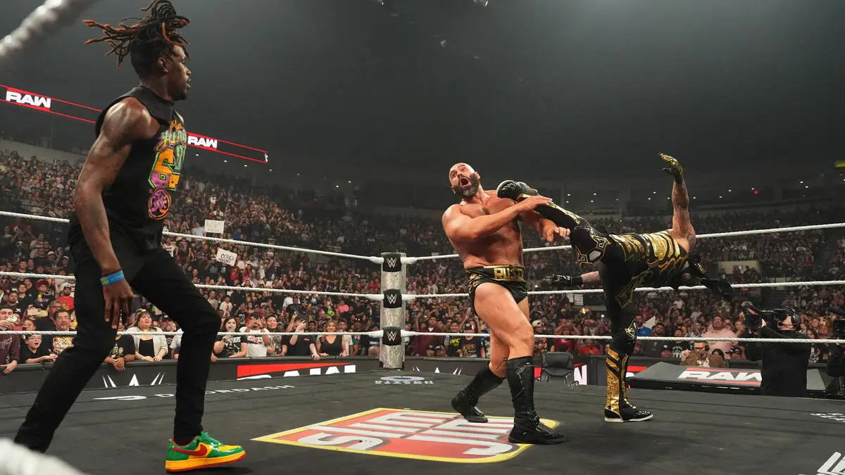 WWE wrestlers in a ring; one man delivers a high kick to a masked opponent while another watches, crowd cheering in the arena.