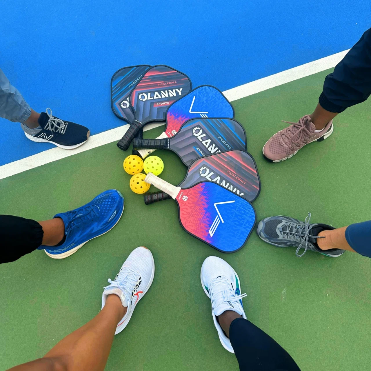 Group of pickleball paddles and bright yellow balls arranged in a circle on a blue-green court, surrounded by players' sneakers.
