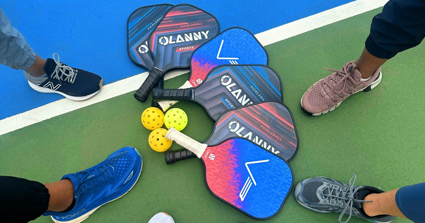 Group of pickleball paddles and bright yellow balls arranged in a circle on a blue-green court, surrounded by players' sneakers.