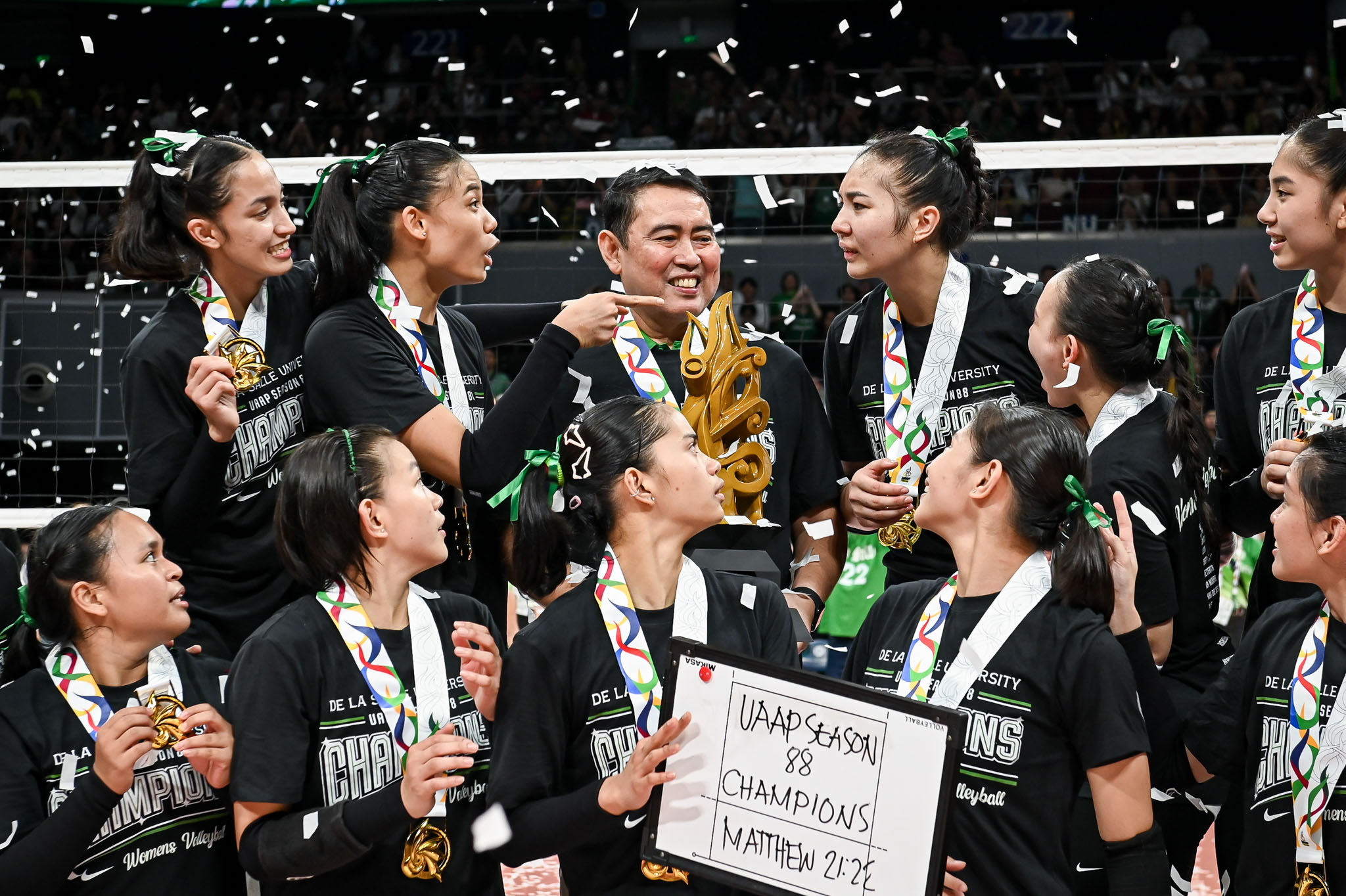 Group of female volleyball players in black jerseys celebrate with a gold trophy and medals as confetti falls.