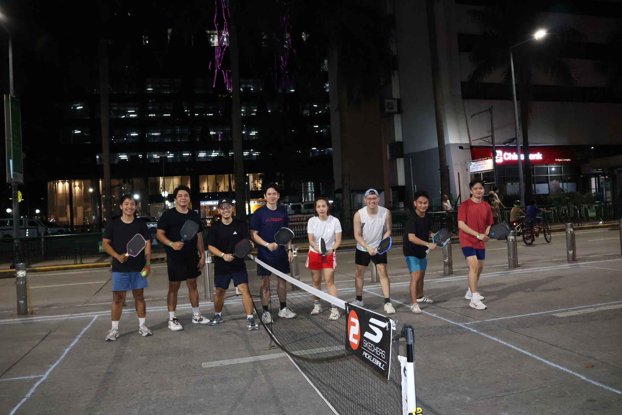 Street-pb-ortigas - the game Group of people standing with pickleball paddles in a parking-lot court at night, a barrier net in front and city lights behind them.