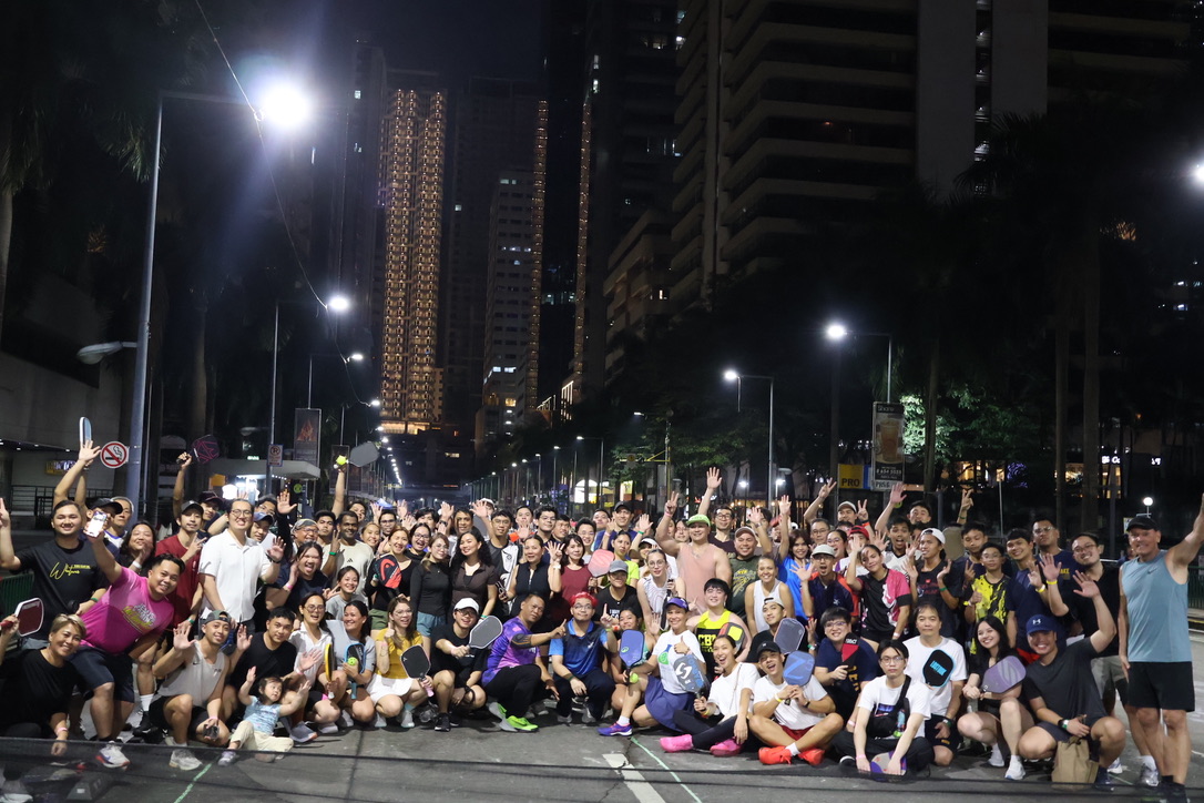 Large group of people posing for a group photo on a city street at night, holding paddles and cheering under streetlights with tall buildings in the background