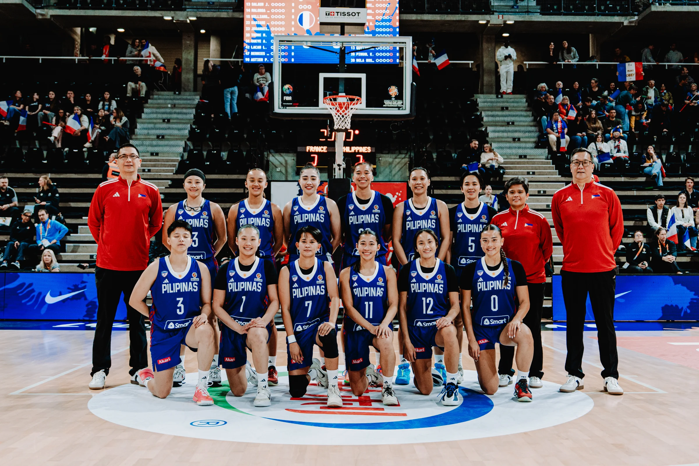 Philippines women's basketball team posing with coaches on a stadium court, crowd in stands behind them.