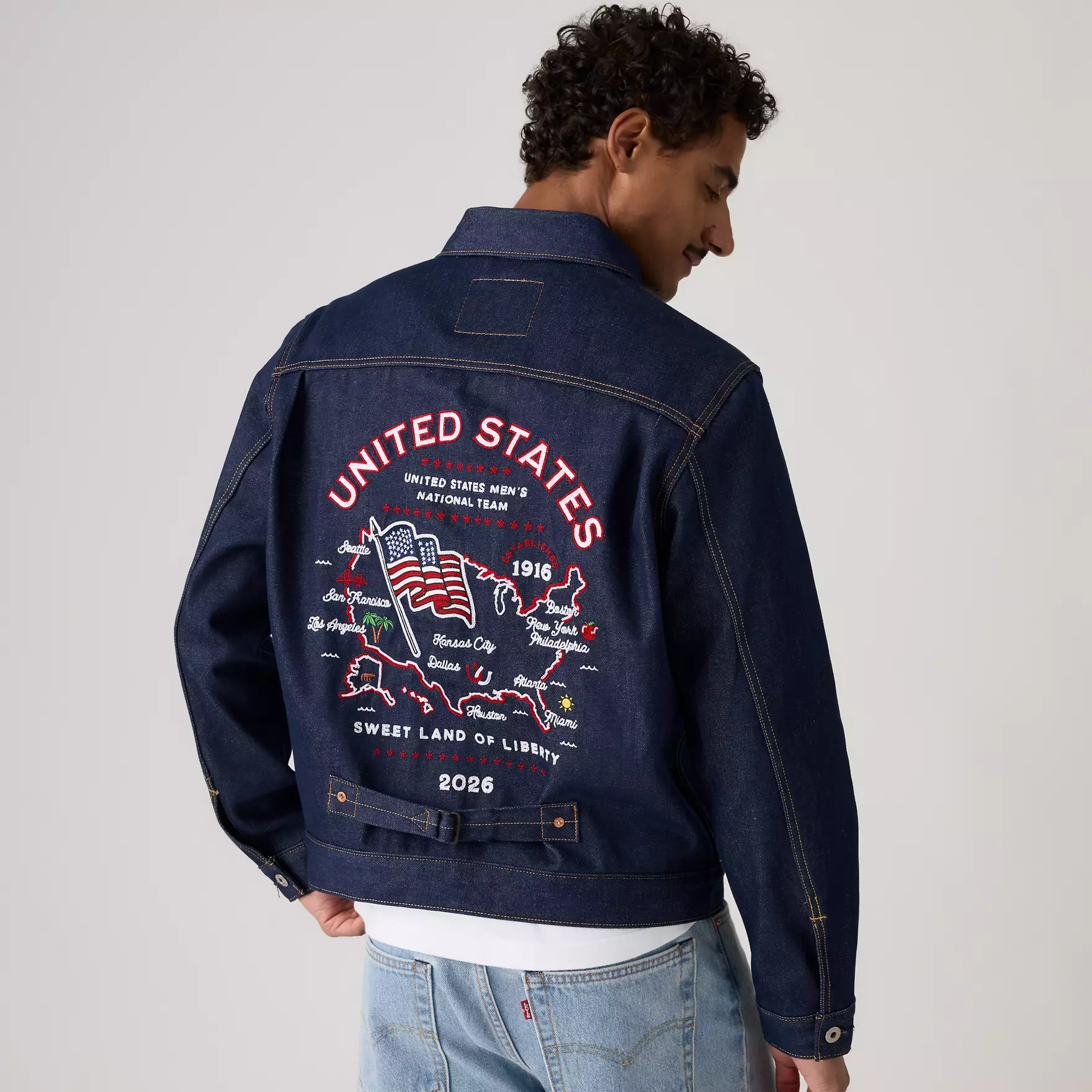 Back view of a man wearing a dark denim jacket with large red and white 'UNITED STATES' embroidery and a U.S. map graphic on the back, 'Sweet Land of Liberty' and 2026 text visible.