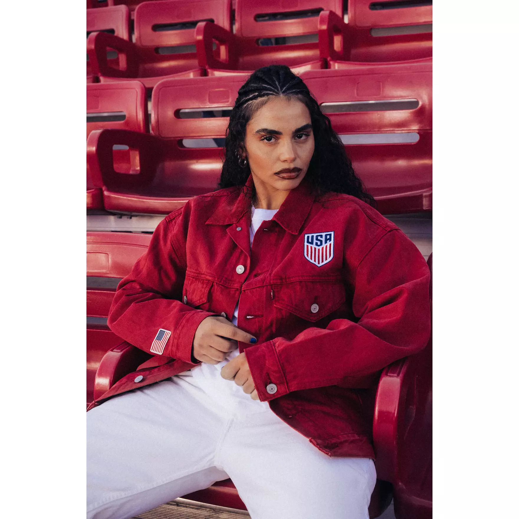 Person seated in a red stadium jacket with USA patch, posing in red bleachers.